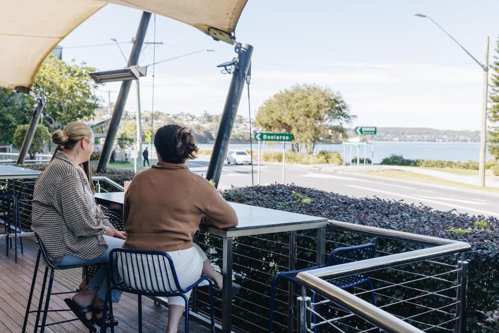 Twee personen zitten aan een buitentafel met uitzicht op een wateroppervlak en een bord dat de richting naar Boolaroo aangeeft.