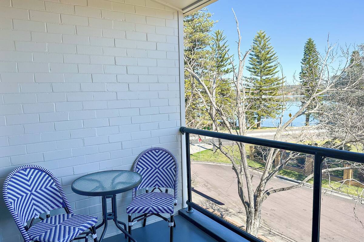 A balcony with two chairs and a small table, overlooking a street and trees.