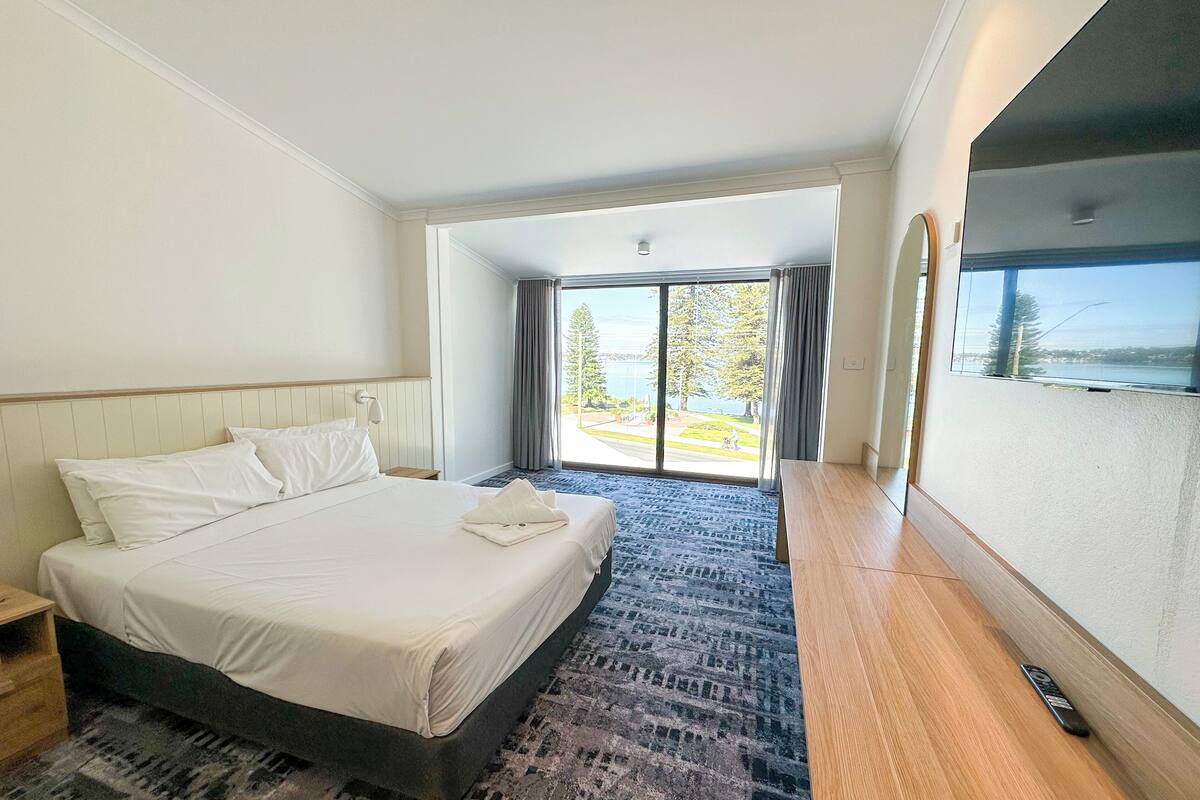 A hotel room with a large bed, a wooden nightstand, a desk, and a view of a body of water through a large window.