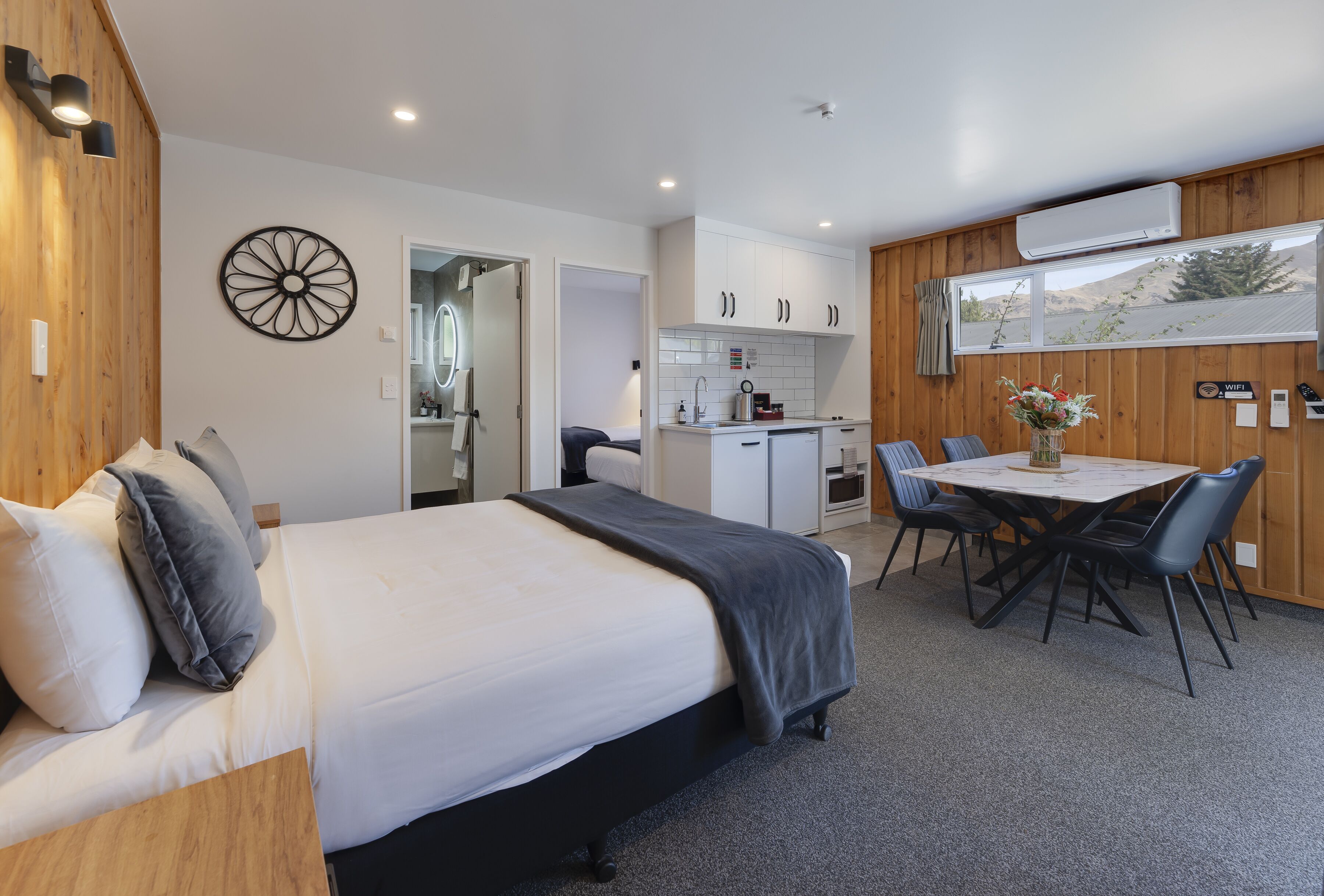 A modern bedroom with a large bed, a dining table with chairs, a kitchen area, and a view of the outdoors.