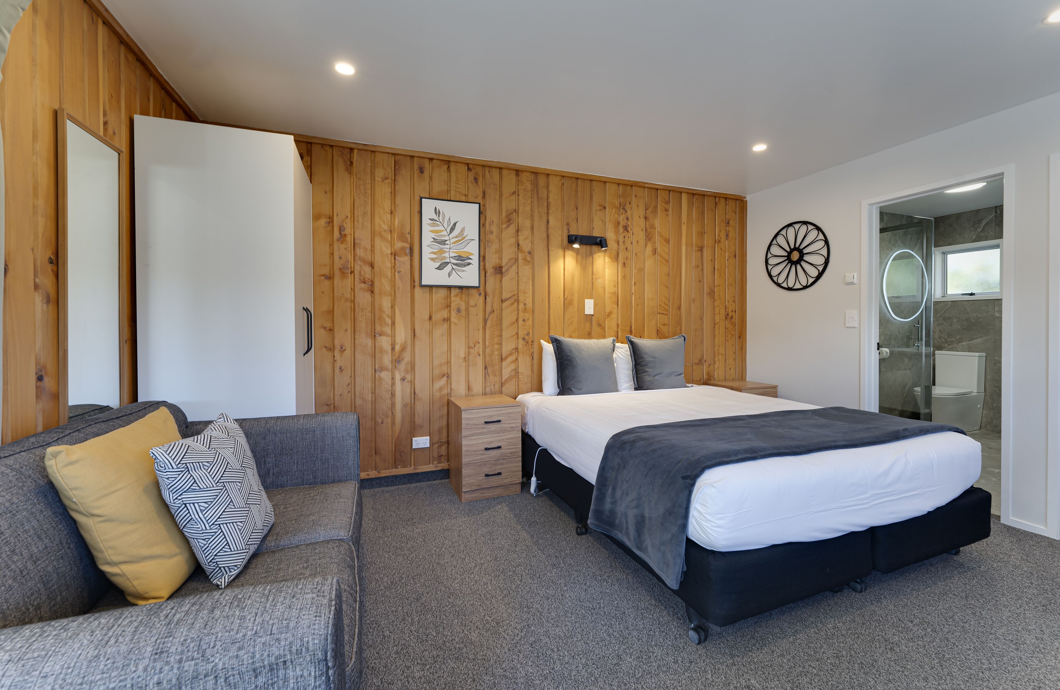 A modern bedroom with a wooden wall panel, a bed with a black headboard, a grey sofa with patterned cushions, and a bedside table.