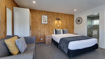 A modern bedroom with a wooden wall panel, a bed with a black headboard, a grey sofa with patterned cushions, and a bedside table.
