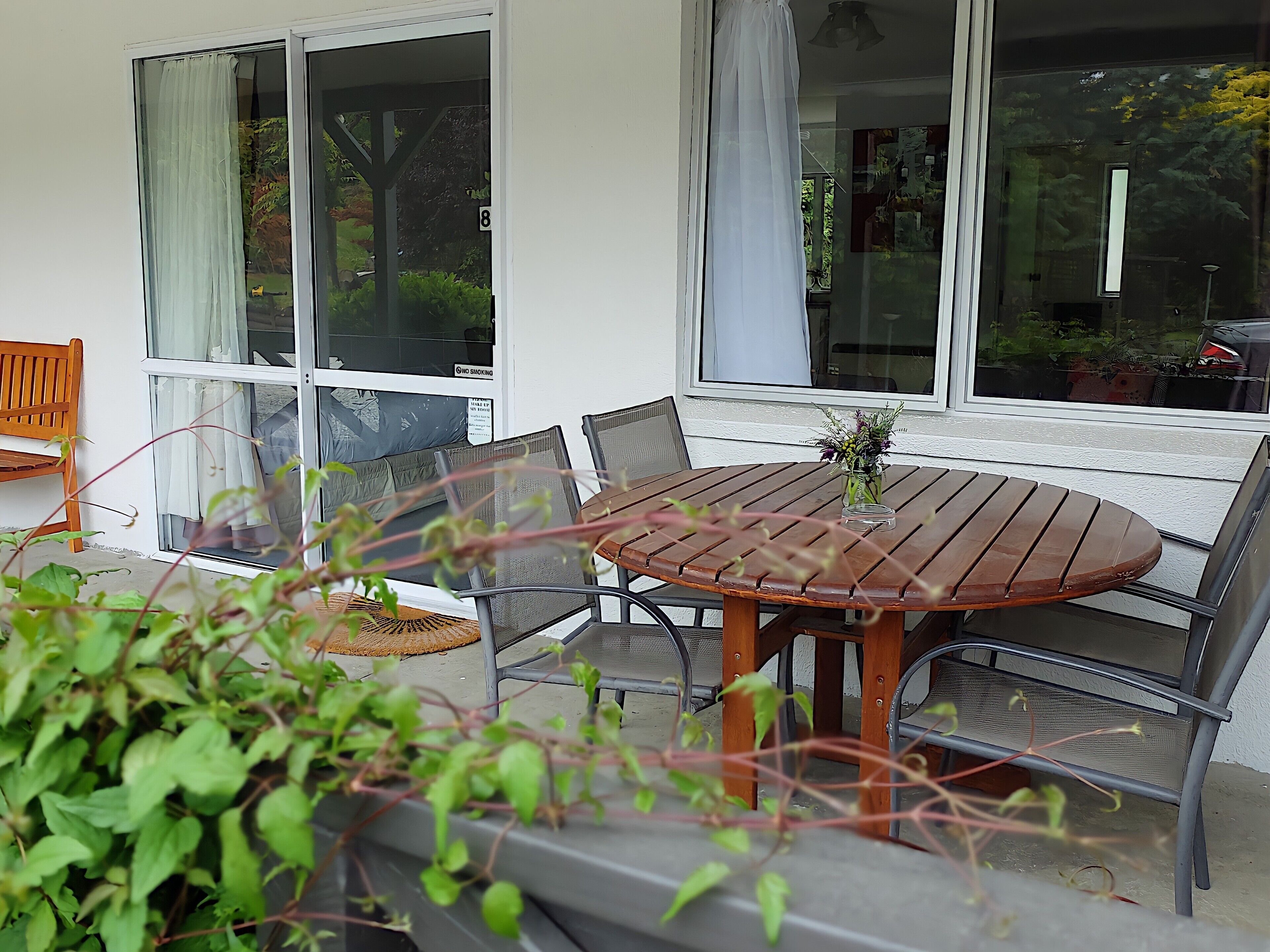 An outdoor seating area with a round wooden table and chairs, a bench, and a glass door leading to a garden.