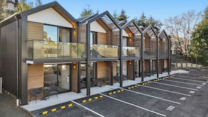 Un hotel moderno con una hilera de habitaciones que tienen balcones, mobiliario exterior y un estacionamiento.