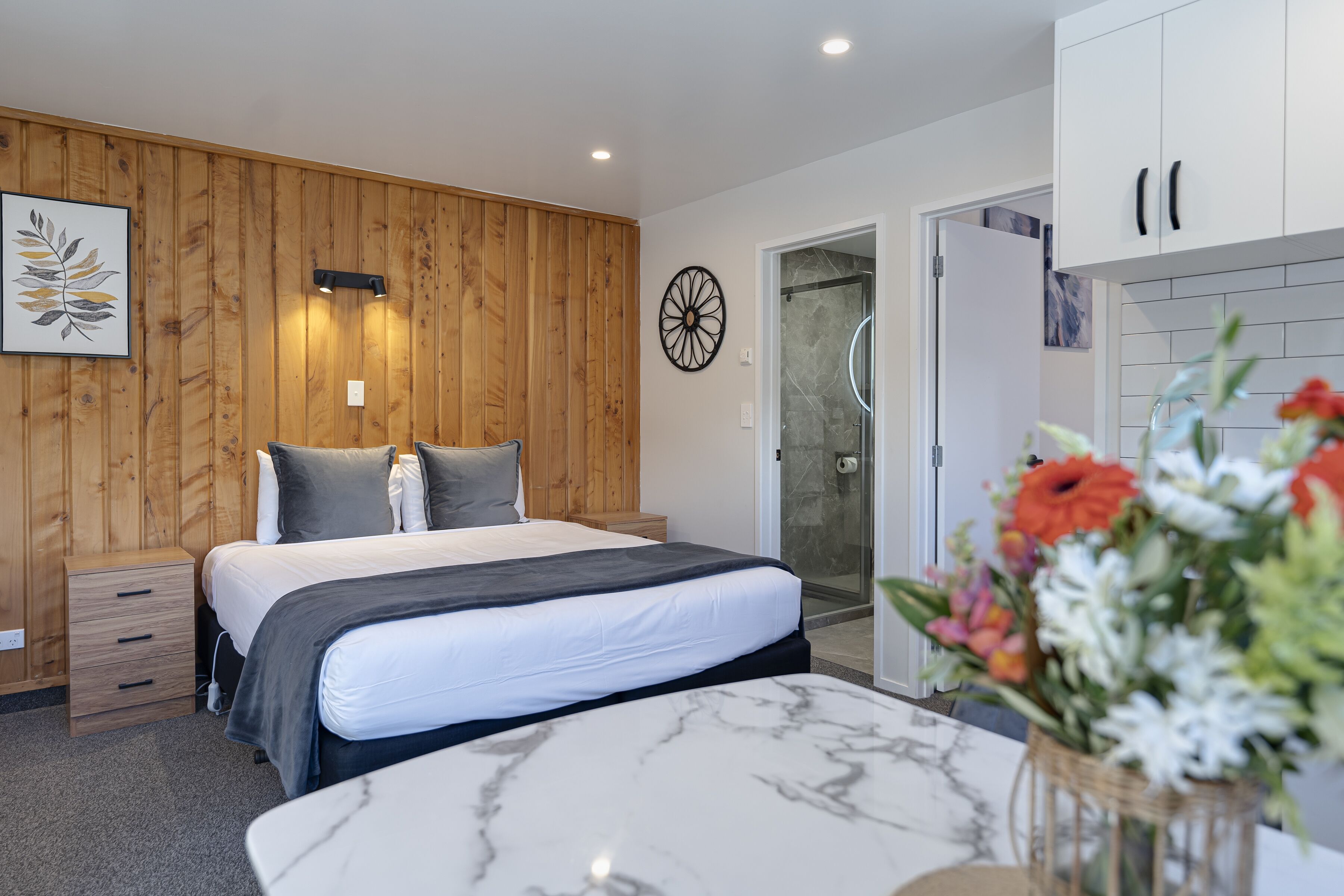 A bedroom with a bed, bedside tables, a wooden wall panel, a shower area, and a bathroom with a sink and mirror.