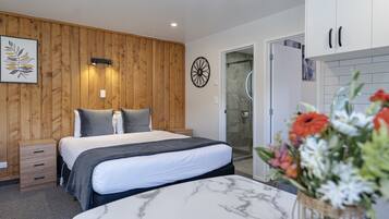 A bedroom with a bed, bedside tables, a wooden wall panel, a shower area, and a bathroom with a sink and mirror.