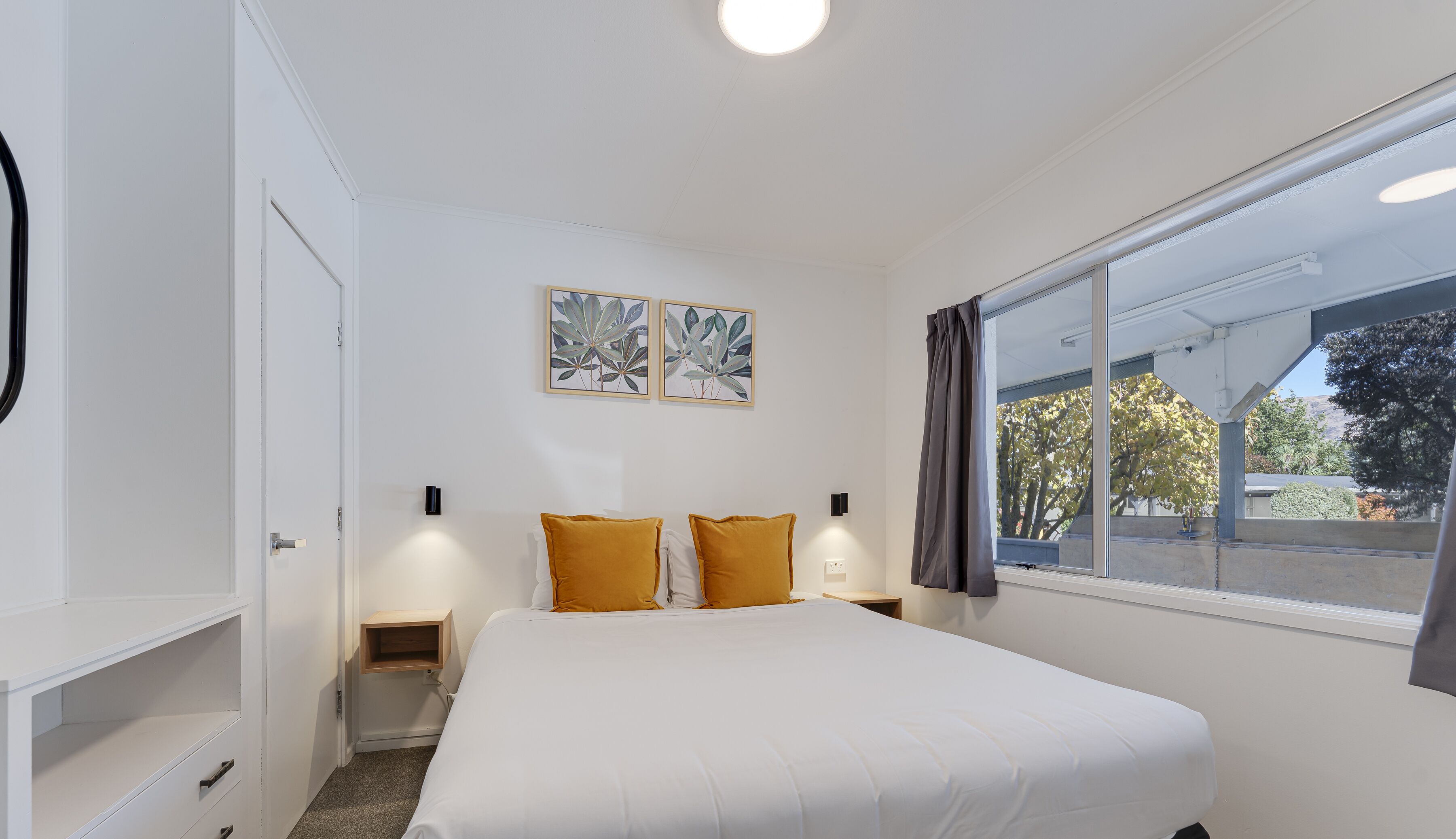 A bedroom with a large bed, two wall-mounted nightstands, a bedside table, a window with curtains, and two wall-mounted wall lights.