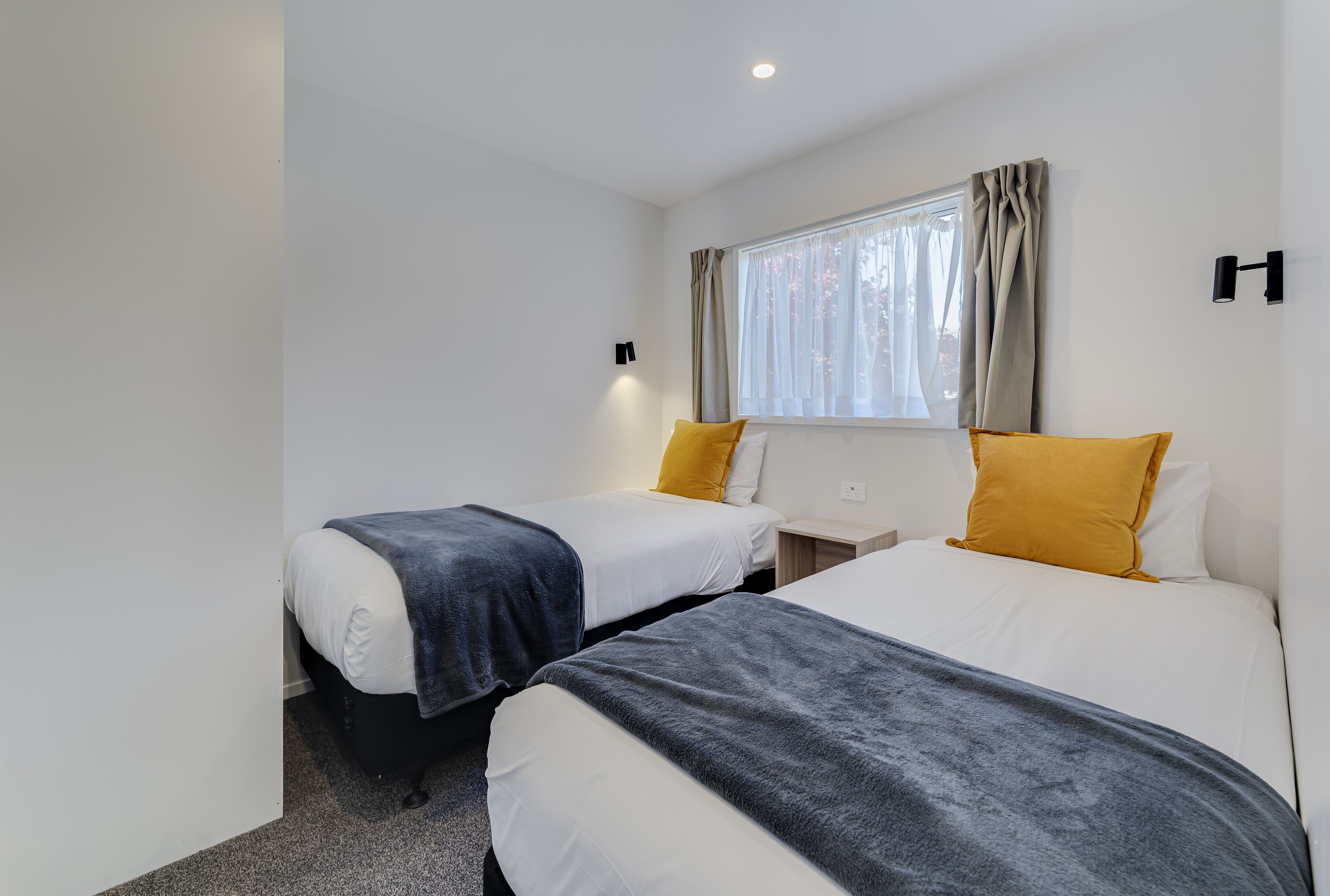 A hotel room with two beds, a window with curtains, a nightstand, and a wall-mounted light fixture.