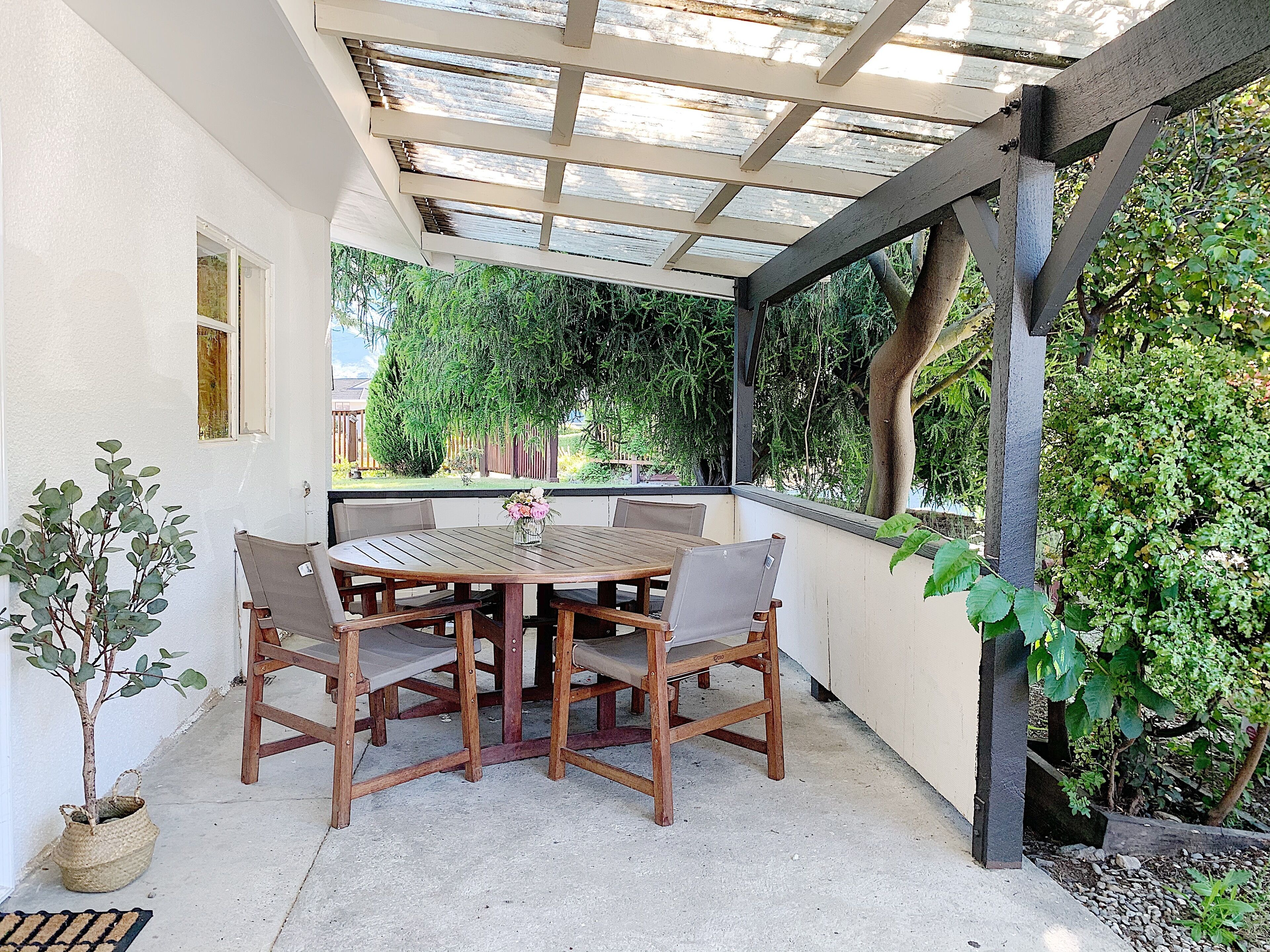 A covered patio with a wooden table and chairs, surrounded by greenery.