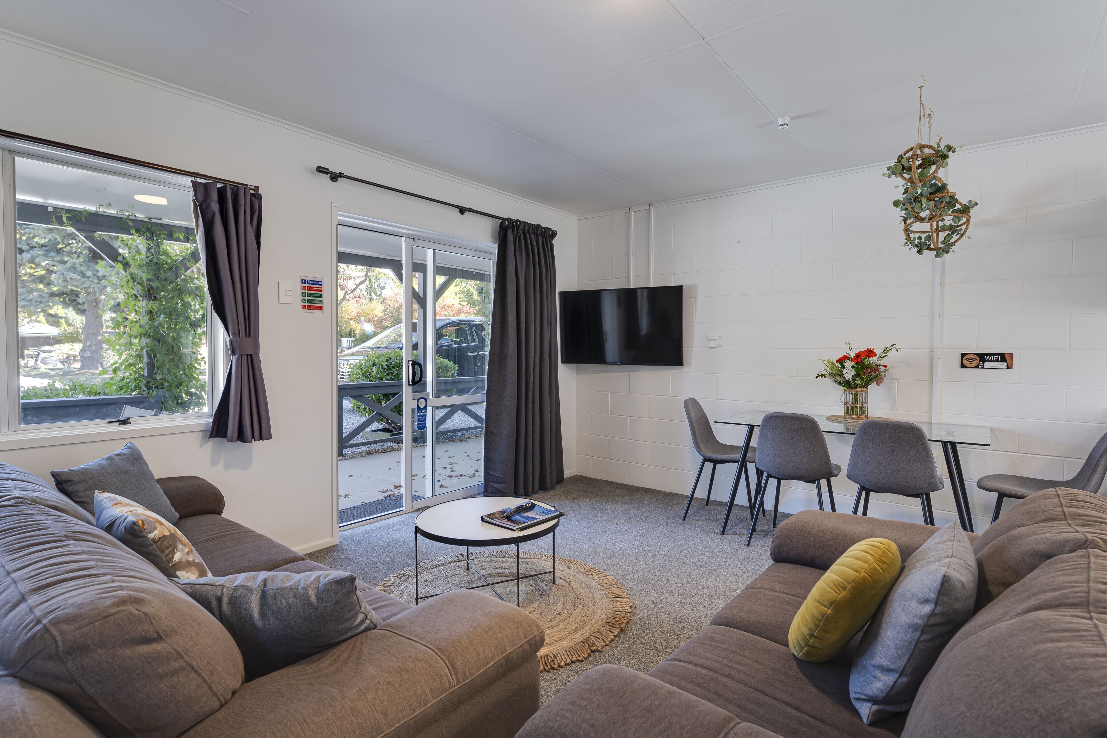 A modern living room with a sofa, a small table, and a TV mounted on the wall. There is a sliding glass door leading to an outdoor area.
