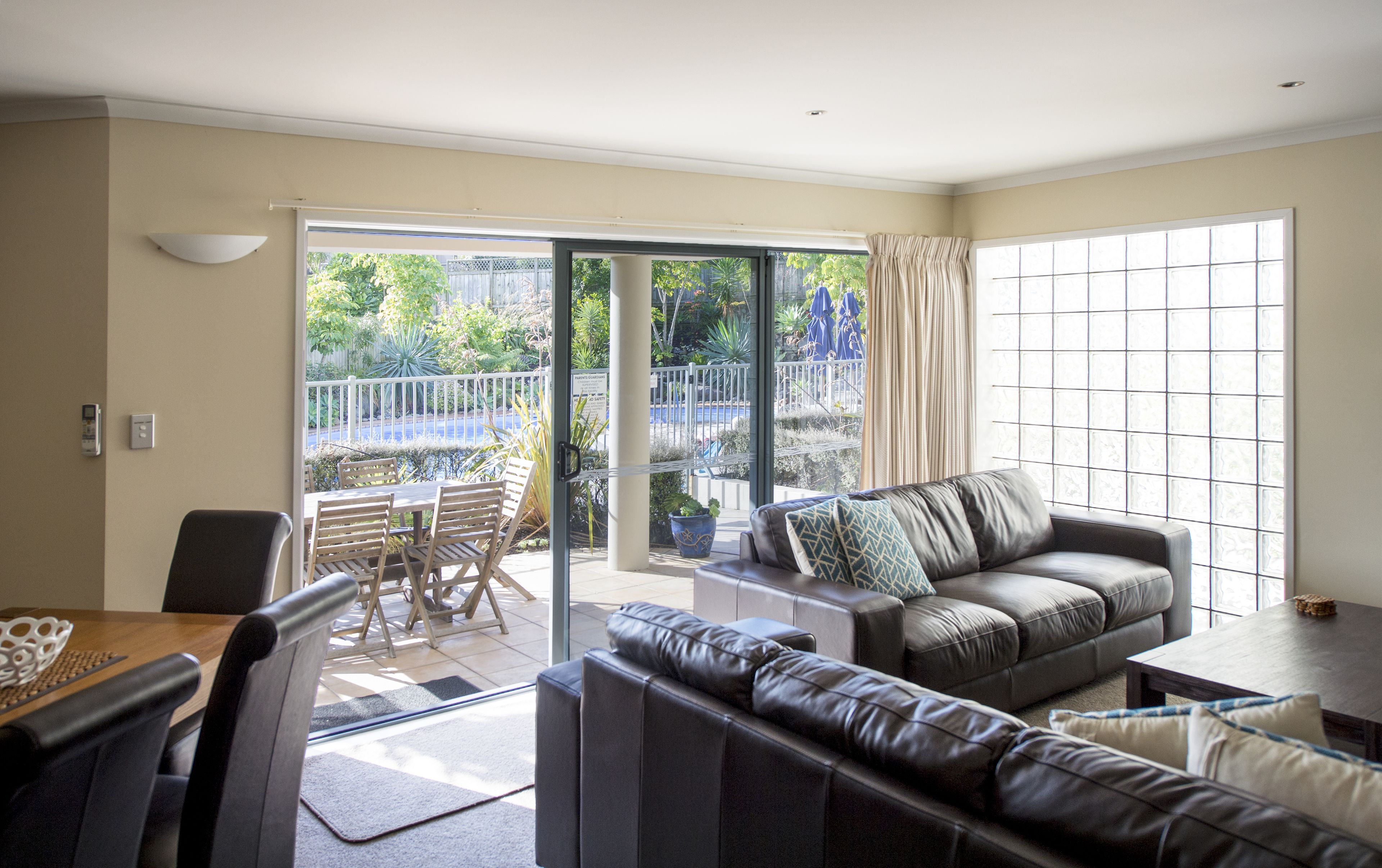 A living room with a sofa, a dining table with chairs, and a sliding glass door leading to an outdoor area with a view of greenery and a fence.