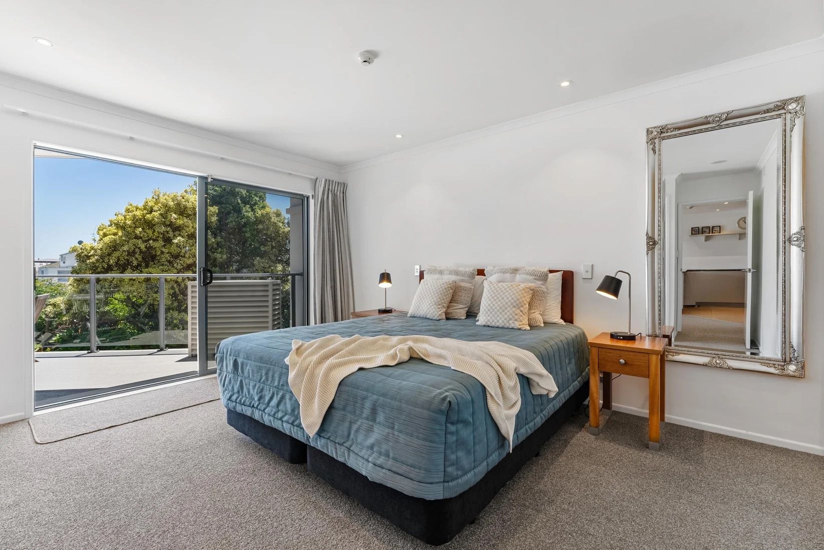 A bedroom with a large bed, a nightstand, a mirror, and a sliding glass door leading to a balcony.