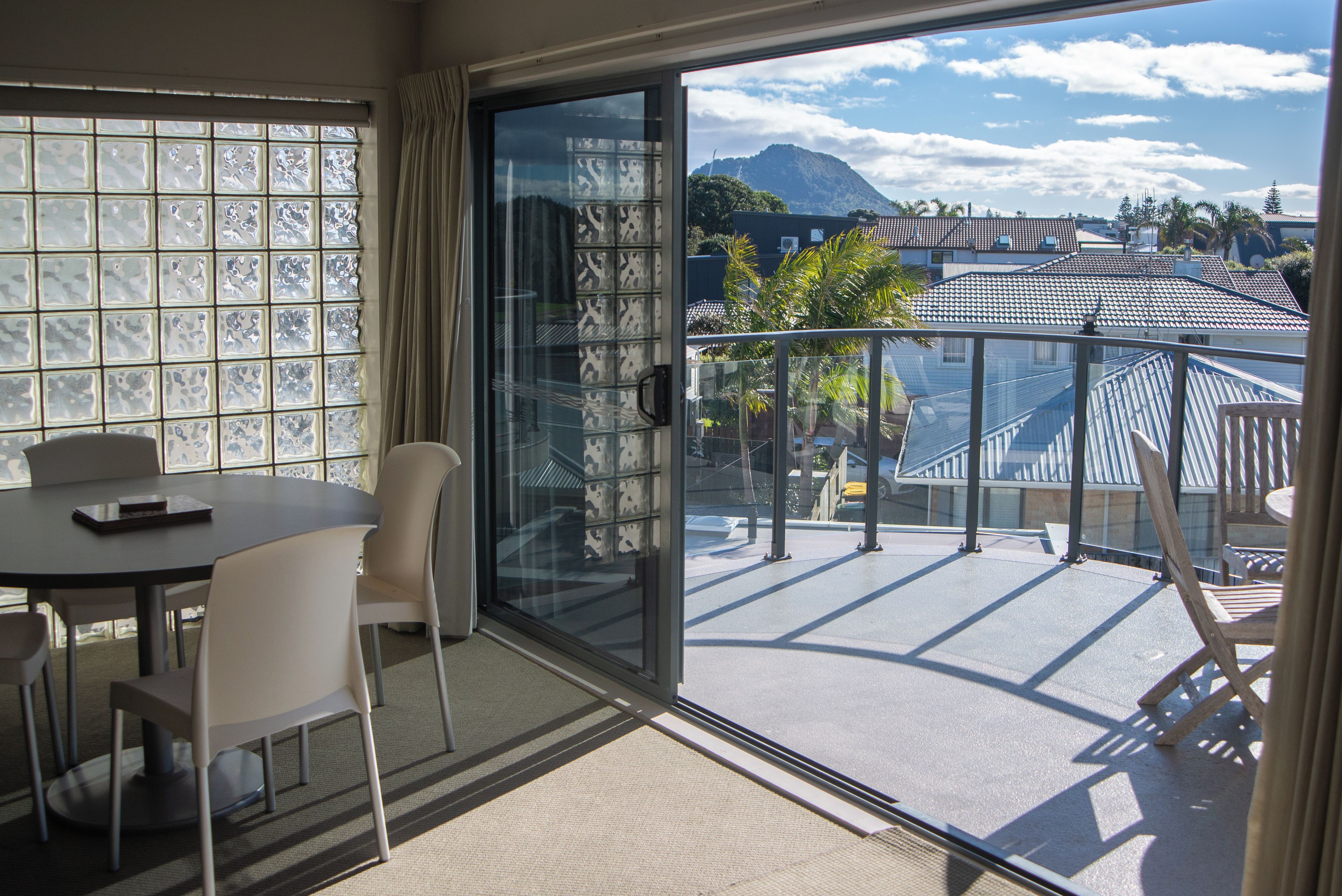 Una habitación con una mesa y sillas, una puerta corrediza de vidrio que da a un balcón con vistas a edificios y una montaña.