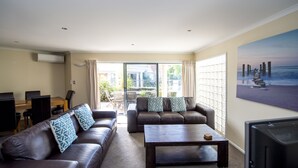 A living room with a brown leather sofa, a wooden coffee table, and a TV. There is a balcony with a view of the ocean and a painting of a beach scene on the wall. - The Reef Beachfront Apartments (Tauranga)