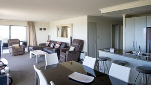 A modern hotel room with a dining area, a living room with a sofa and chairs, a kitchen with a stainless steel refrigerator, and a large window with curtains. - The Reef Beachfront Apartments (Tauranga)