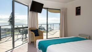 A hotel room with a large bed, a television mounted on the wall, a chair with a cushion, and a balcony with a view of the ocean. - The Reef Beachfront Apartments (Tauranga)