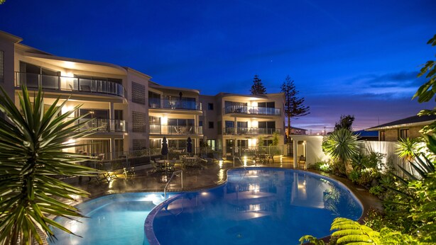 A pool area with a curved pool, lounge chairs, and a building with multiple balconies. - The Reef Beachfront Apartments (Tauranga)