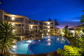 A pool area with a curved pool, lounge chairs, and a building with multiple balconies. - The Reef Beachfront Apartments (Tauranga)