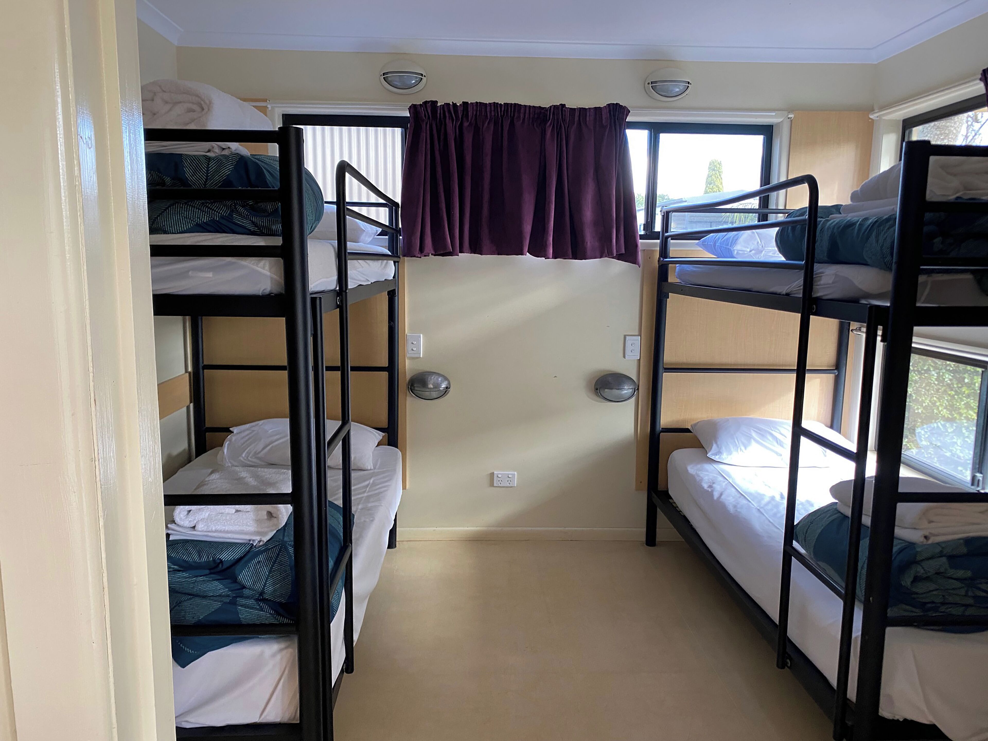 A bunk room with multiple beds, a window with curtains, and a door with a light switch.