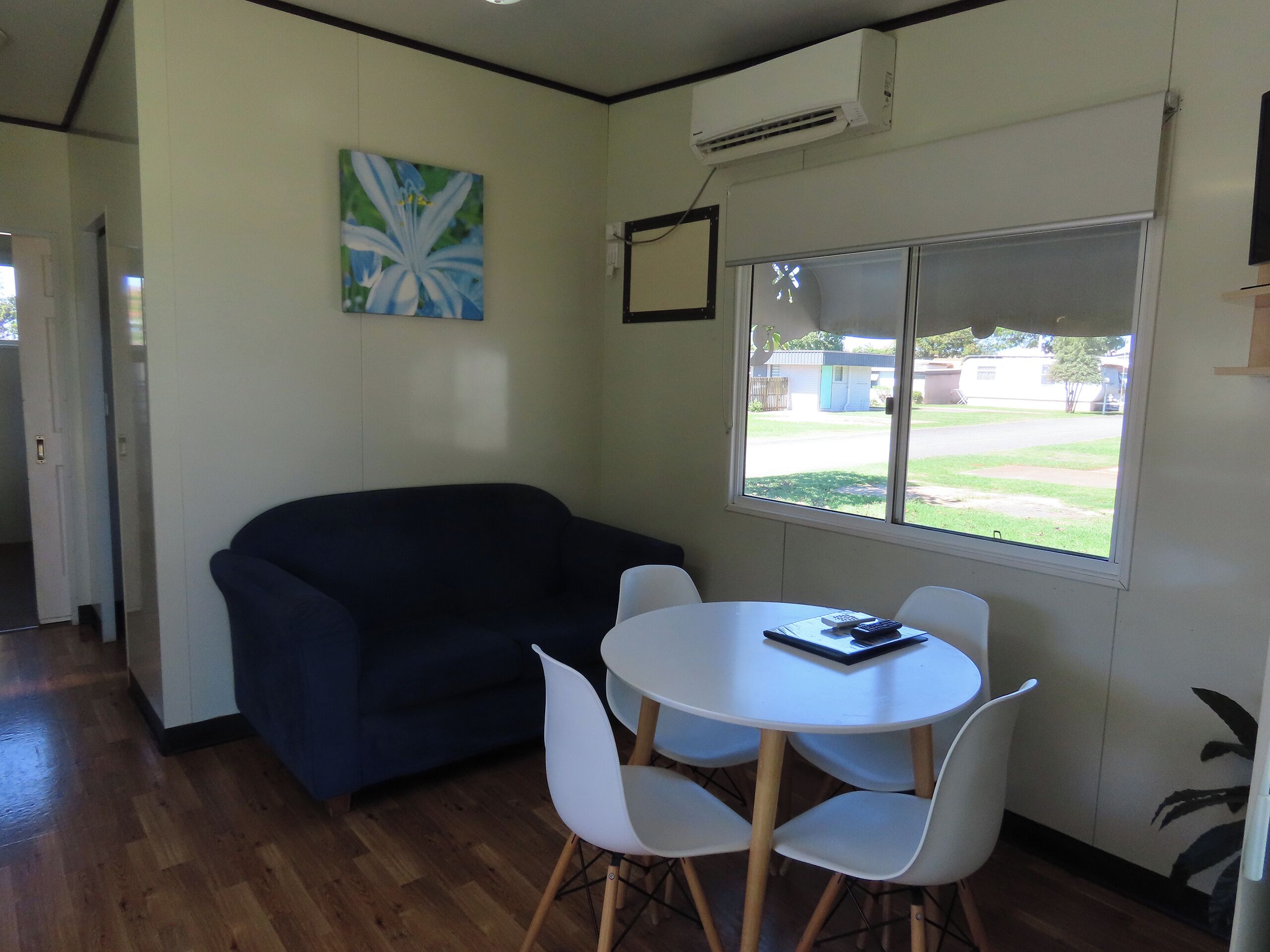 A room with a dark blue sofa, a white round table with four chairs, a large window with a view of a residential area, and an air conditioning unit on the wall.