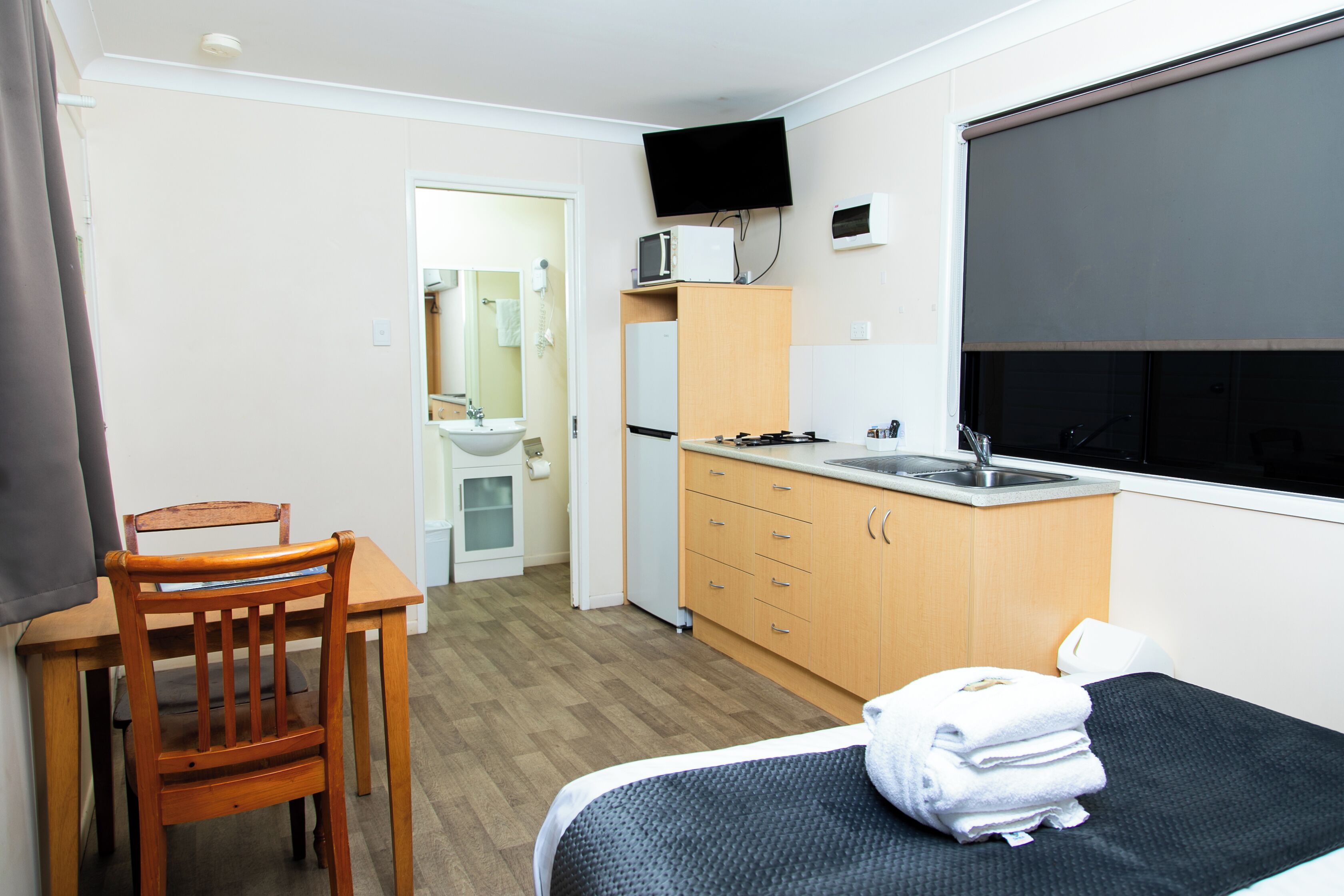 A compact room with a bed, a wooden chair, a small table, a kitchenette, and a TV mounted on the wall.