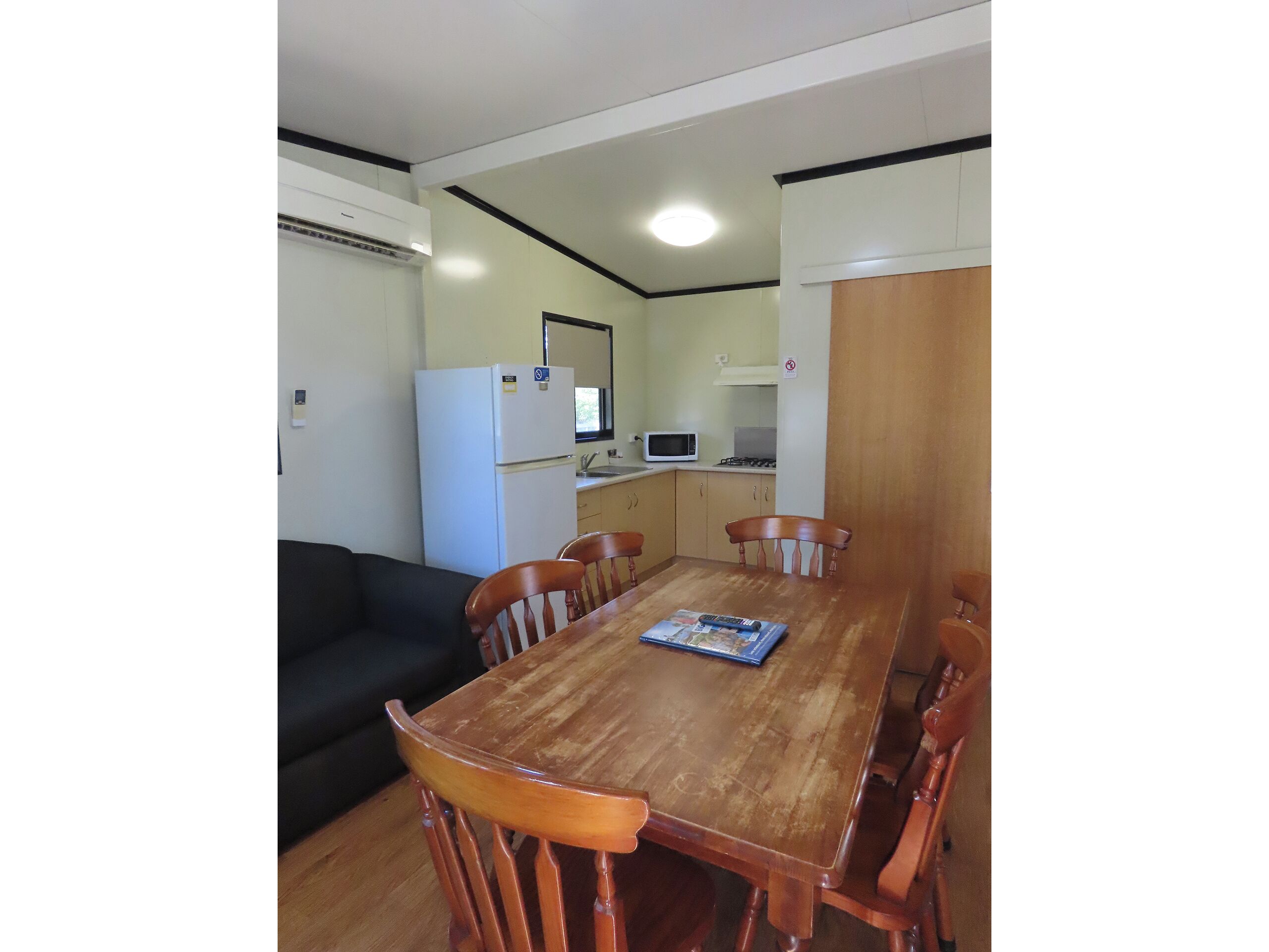 A compact living space with a dining table, wooden chairs, a black sofa, a white refrigerator, a microwave, and a small kitchenette.