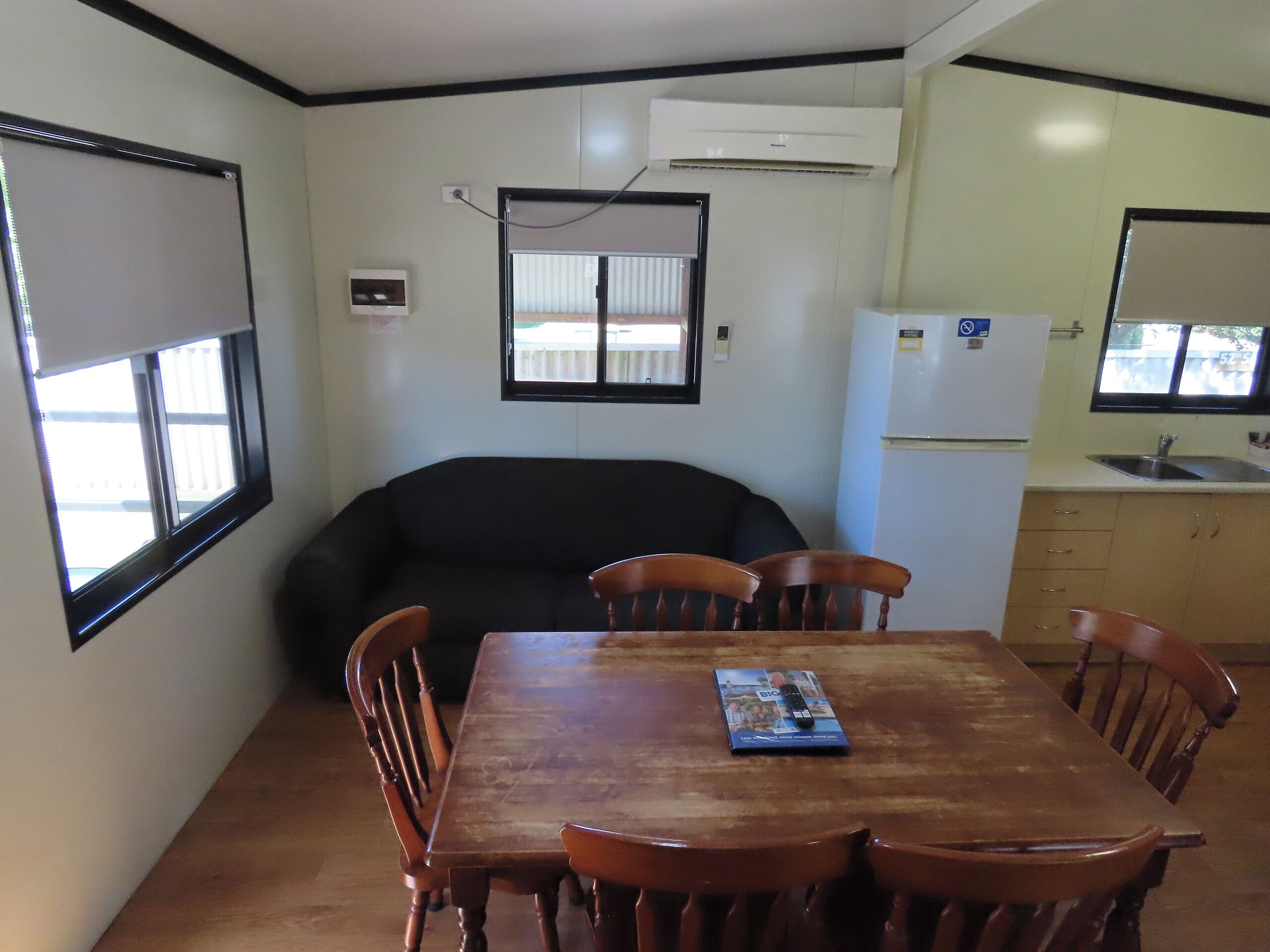 Una habitación compacta con una mesa de comedor de madera, cuatro sillas, un sofá negro, una pequeña kitchenette con refrigerador y pileta, y una ventana con persianas.