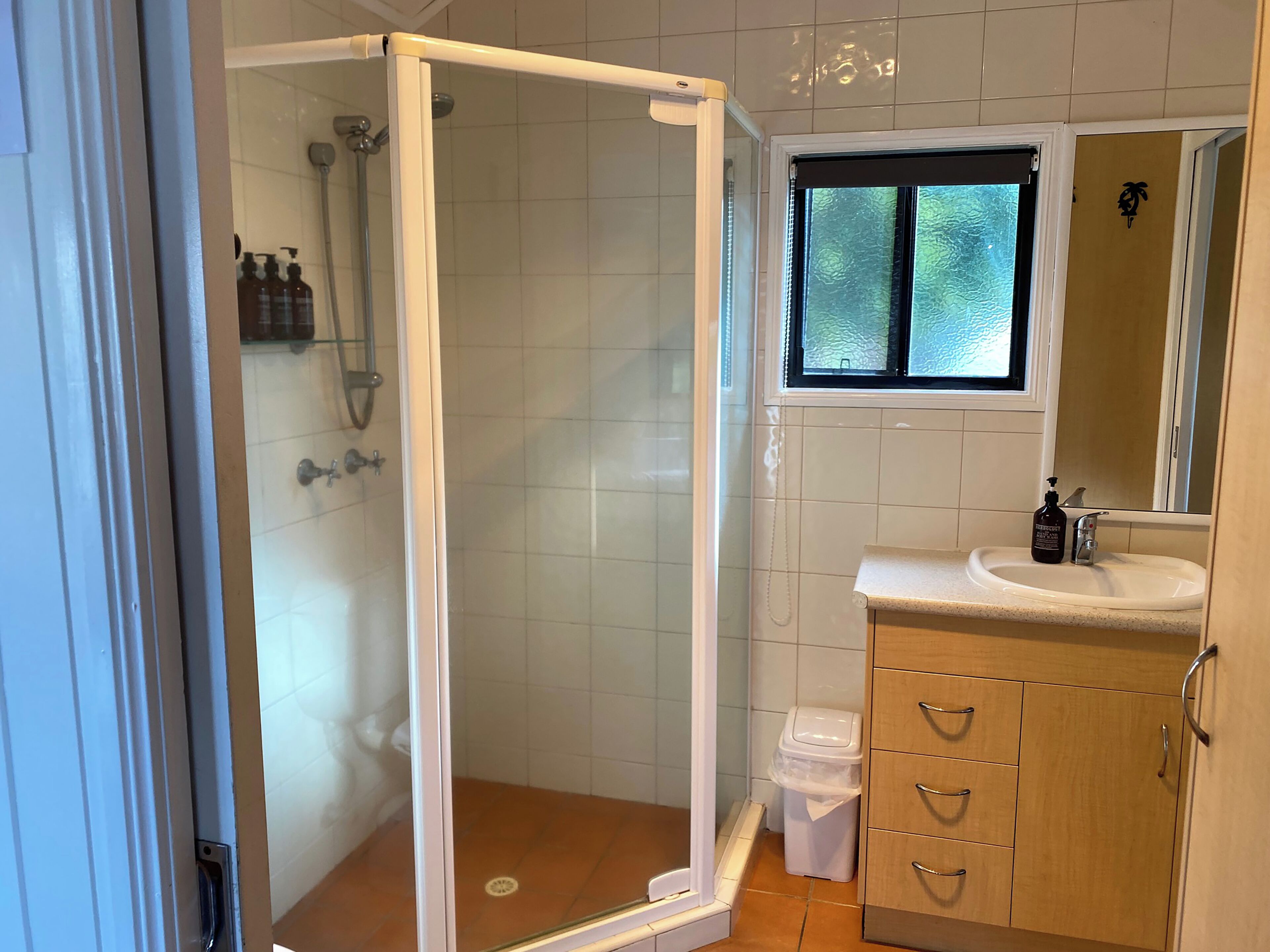 A bathroom with a glass-enclosed shower, a window above the sink, and a wooden cabinet with drawers.