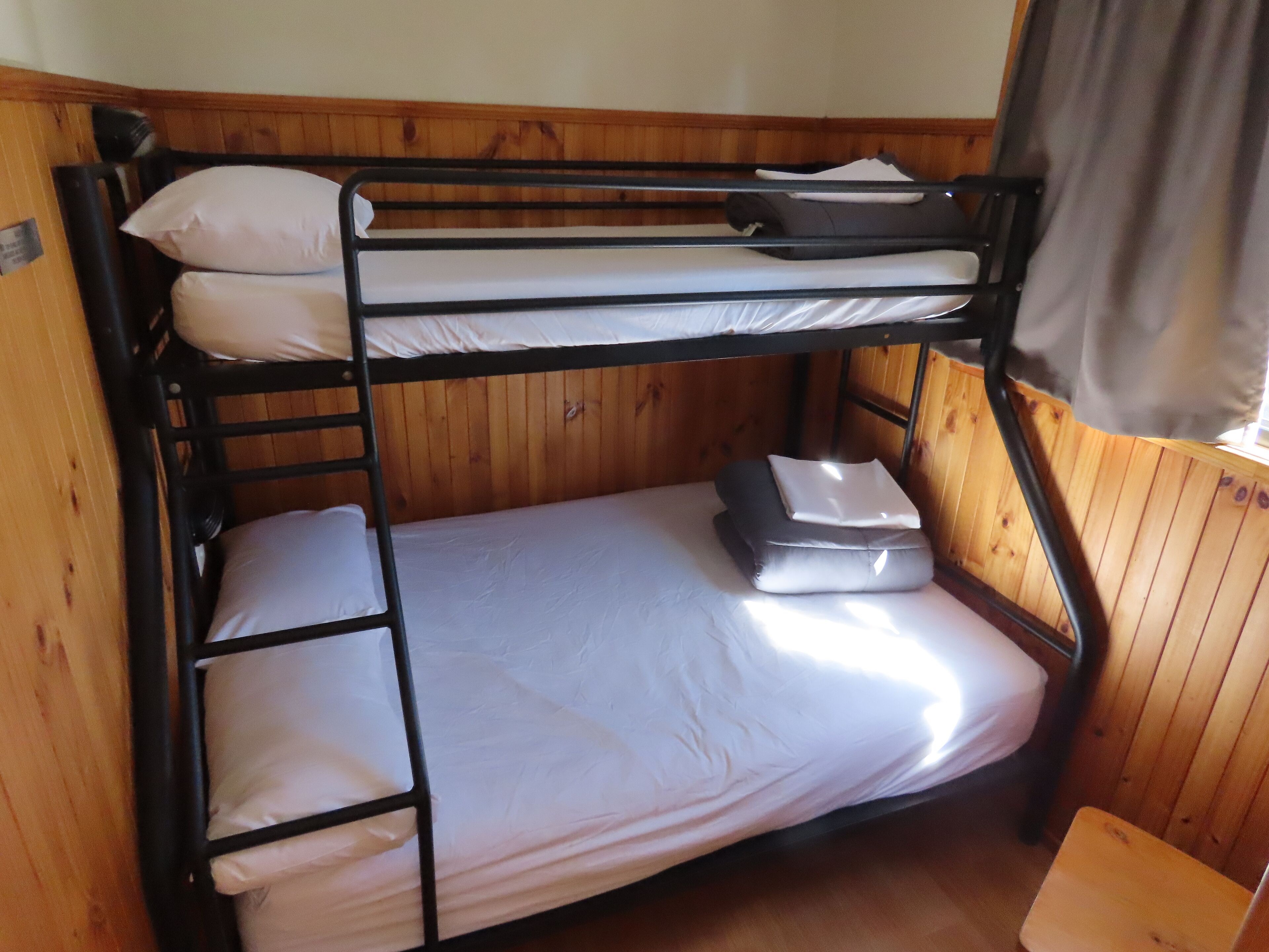 A bunk bed with white bedding and pillows, wooden walls, and a window with curtains.