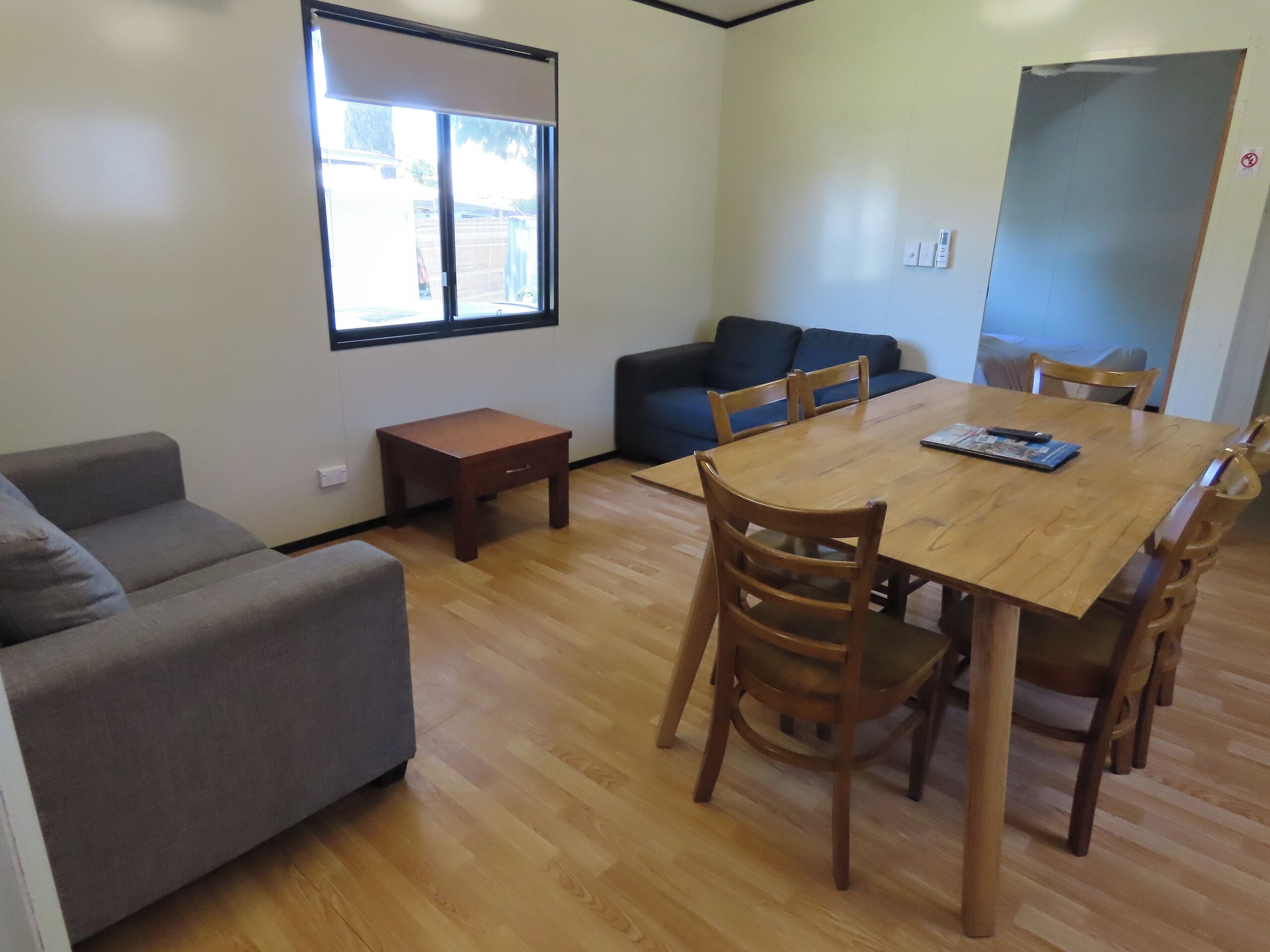 A room with a wooden dining table, four chairs, a grey sofa, a small wooden side table, and a window with blinds.