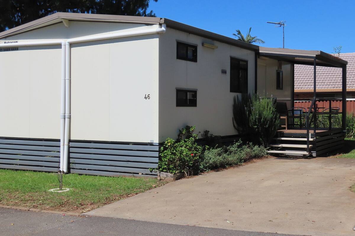 Une maison mobile de plain-pied, avec le numéro « 46 » peint sur le côté, une petite véranda avec une table et des chaises, et un jardin avec des plantes et un petit arbre.