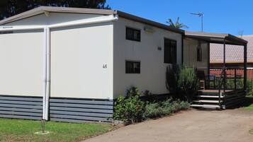 A single-story mobile home with a number '46' on the side, a small porch with a table and chairs, and a garden area with plants and a small tree.