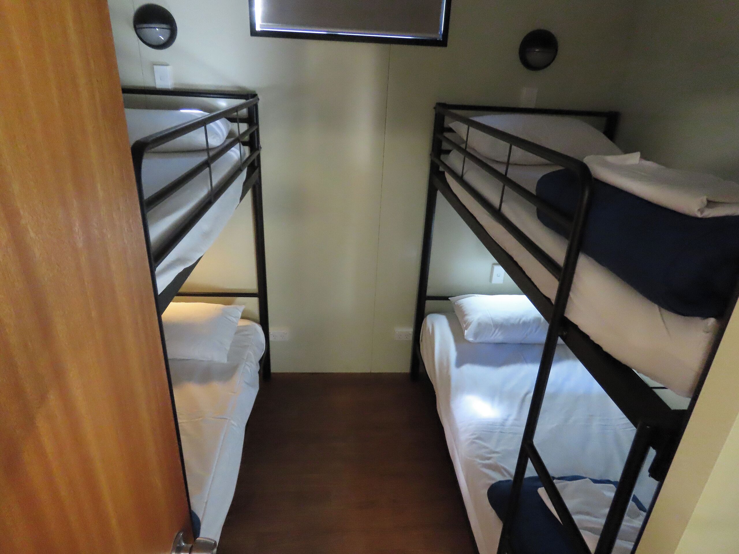 A narrow corridor with bunk beds on either side, a wooden door on the left, and a wall-mounted light fixture.