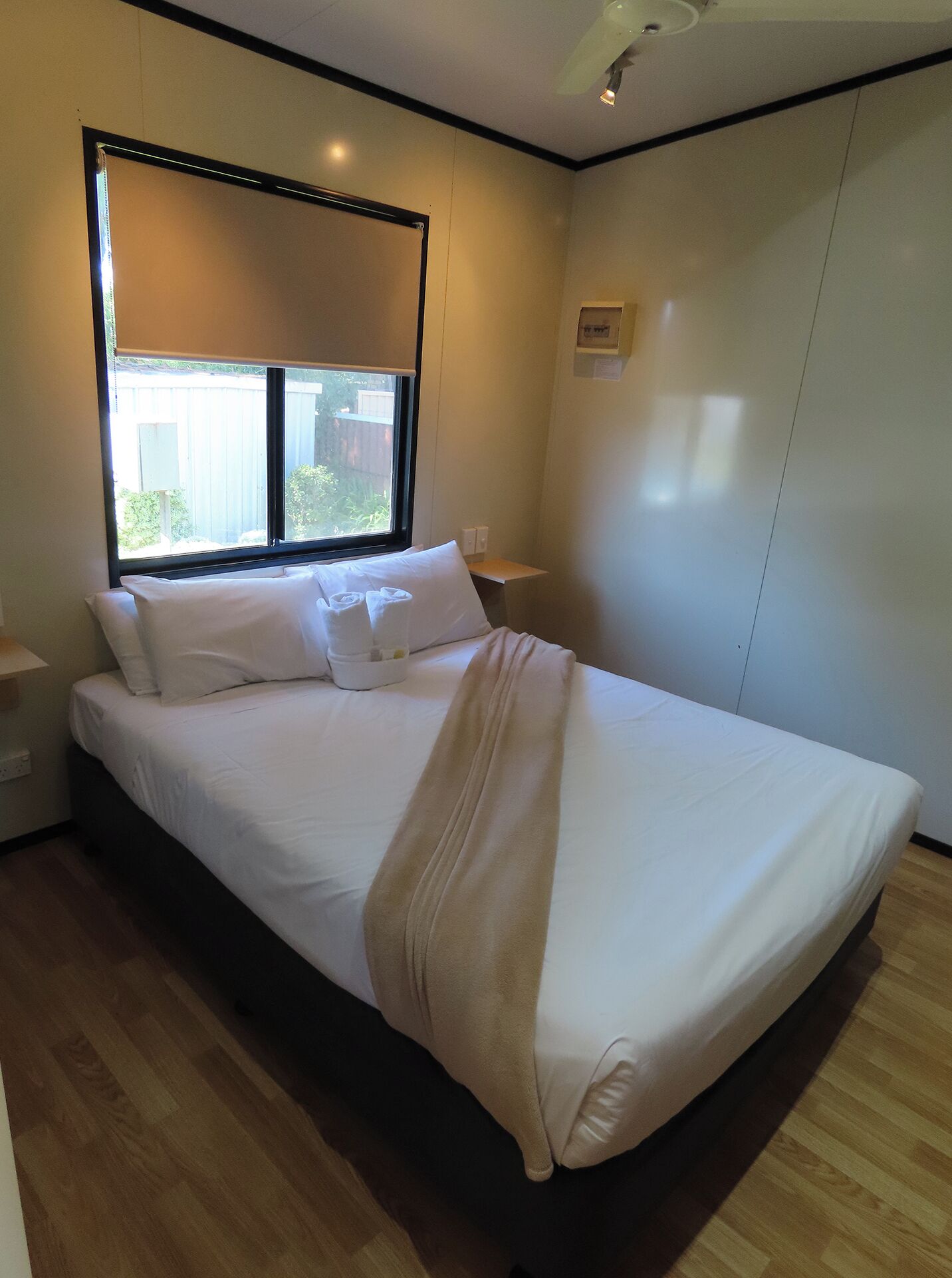 A neatly made bed in a room with a large window, a bedside table, and a wall-mounted light fixture.