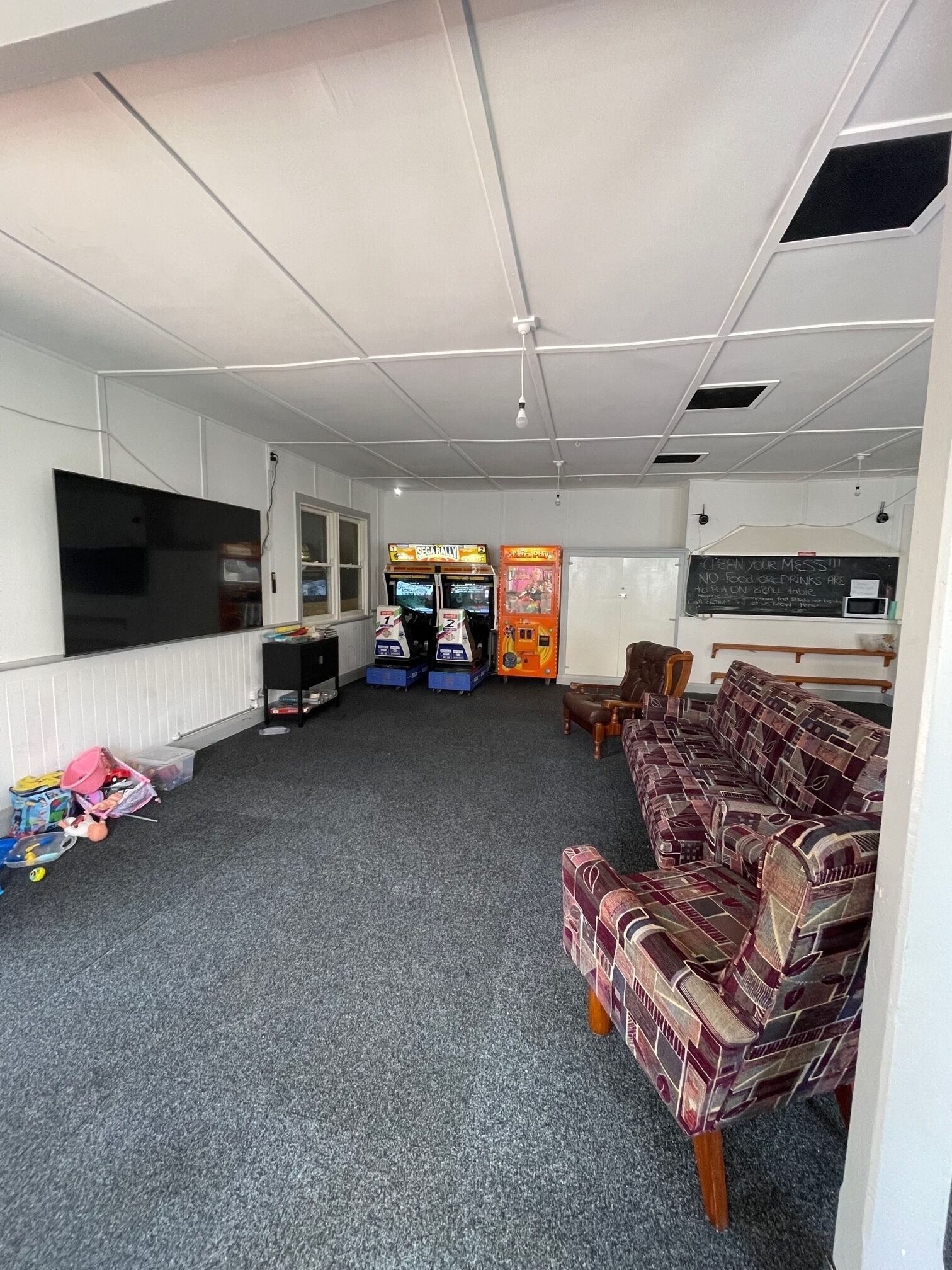 A room with a large flat-screen TV, two arcade machines, a sofa, and a chalkboard with a message.