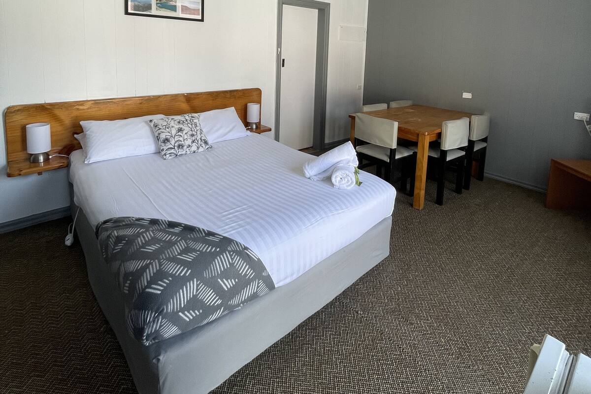 A hotel room with a large bed, a wooden headboard, a desk, and a chair. There is a framed picture on the wall and a door in the background.