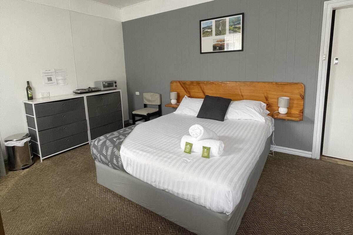 A hotel room with a large bed, a wooden headboard, a bedside table with a lamp, a small desk with a computer, and a trash bin.