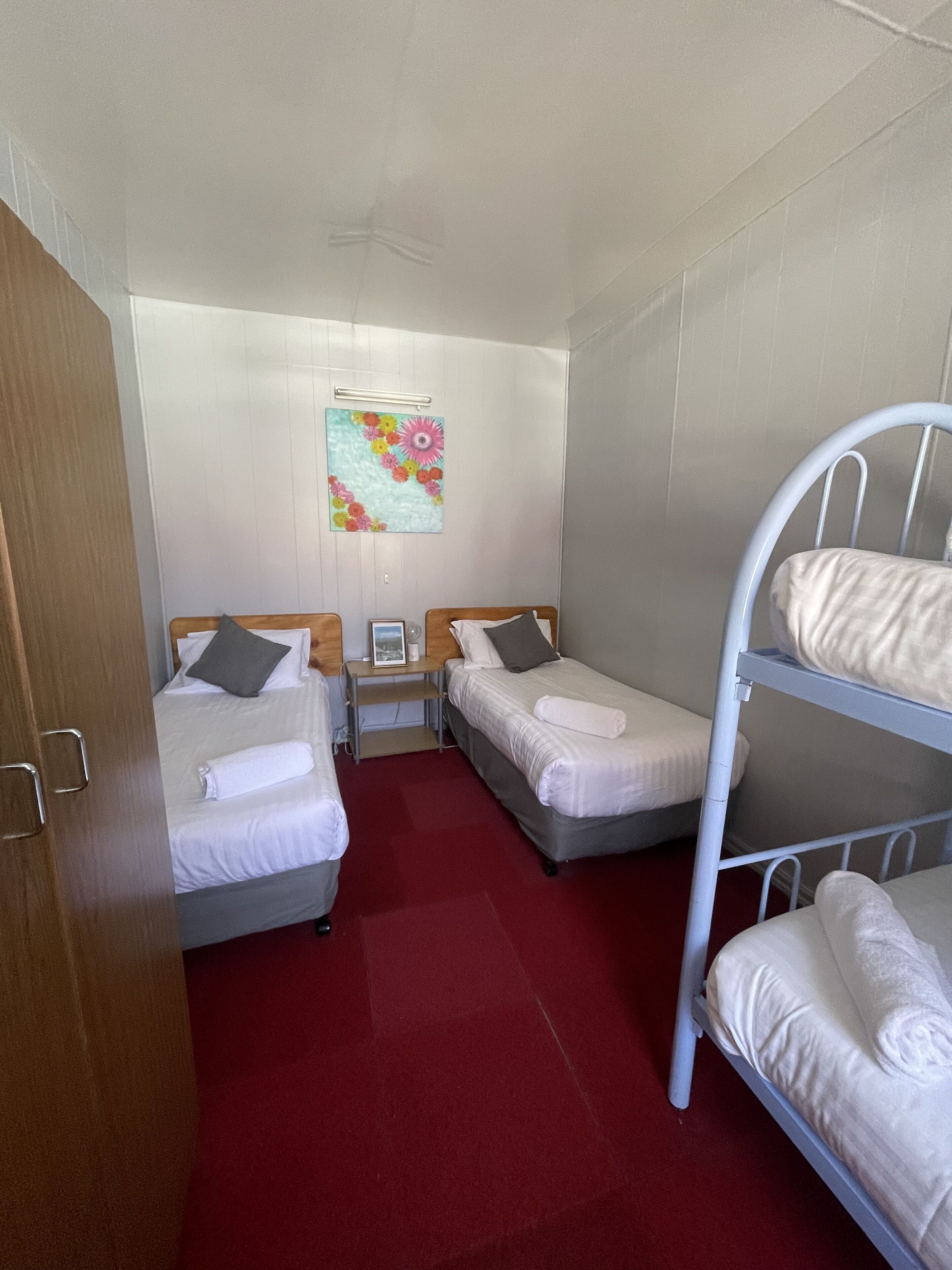 A compact hotel room with two single beds, a wooden wardrobe, a small table with a picture frame, and a colorful floral artwork on the wall.
