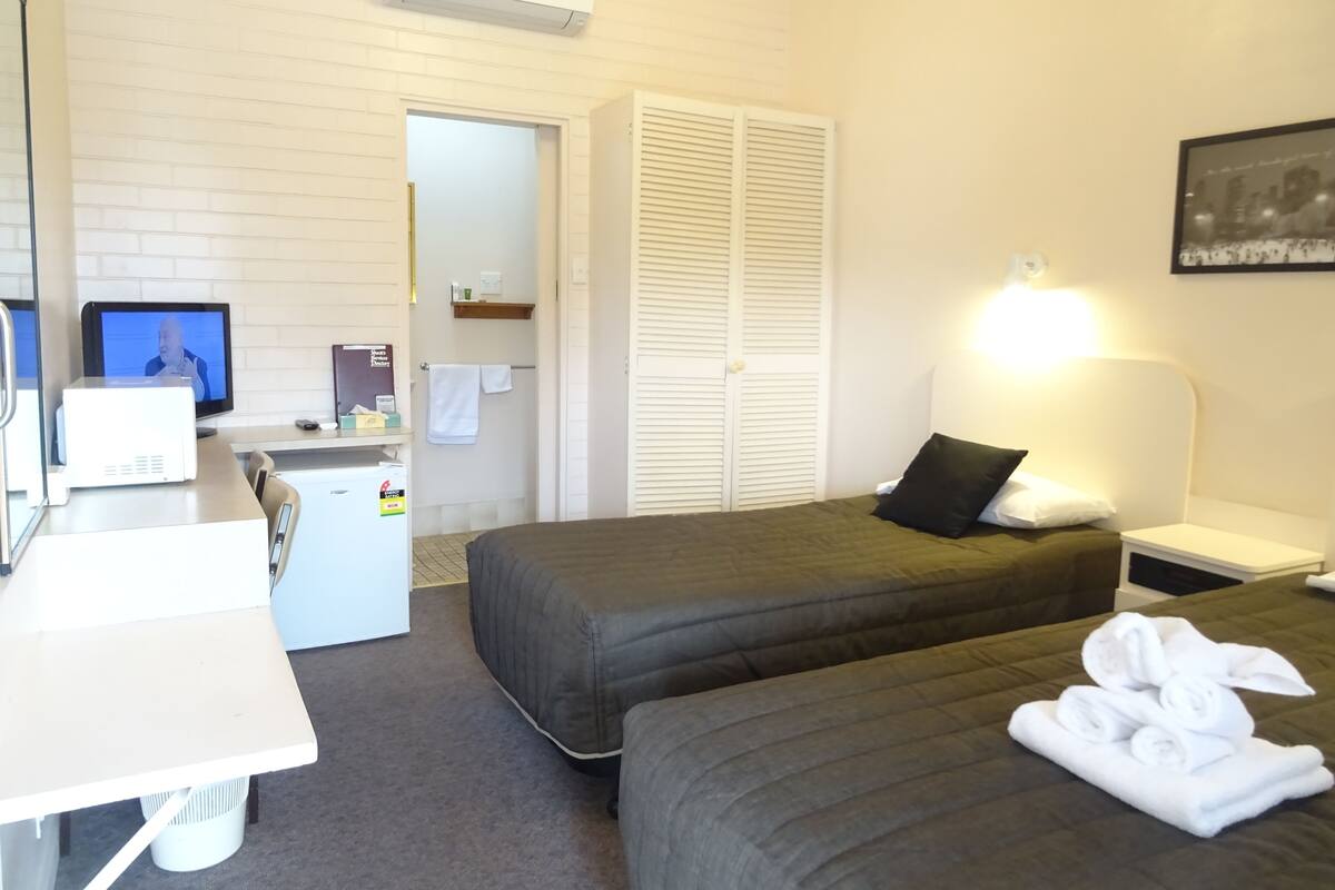 A hotel room with two beds, a desk with a computer, a small fridge, and a white air conditioner.