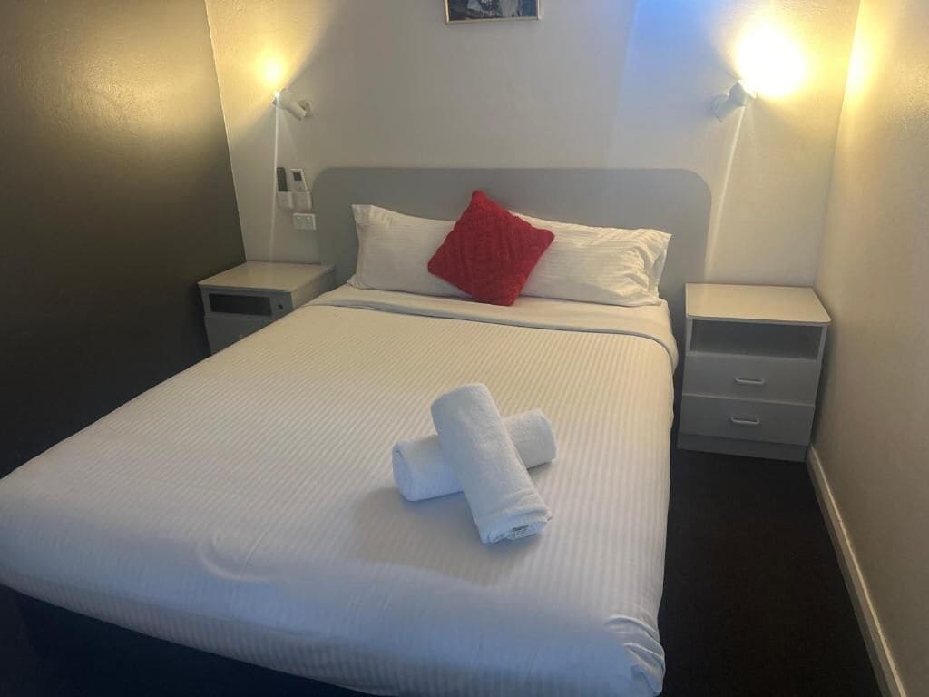 A neatly made bed with white linens and a red pillow, two bedside tables, and a wall-mounted light fixture.