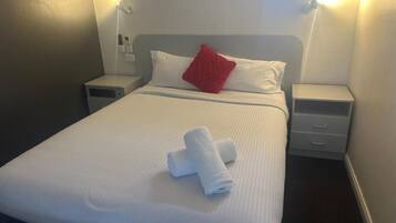 A neatly made bed with white linens and a red pillow, two bedside tables, and a wall-mounted light fixture.