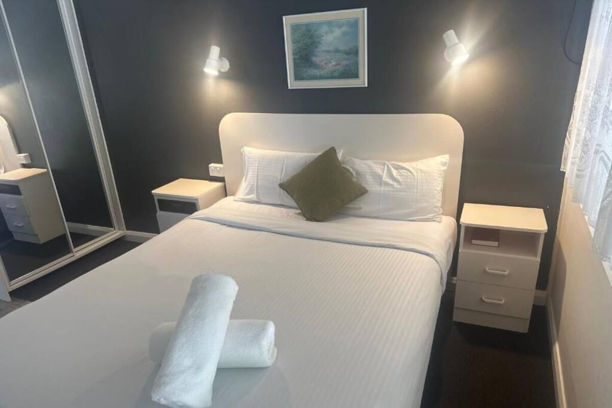 A neatly made bed with white linens and a folded towel, a green pillow, and a framed picture on the wall.