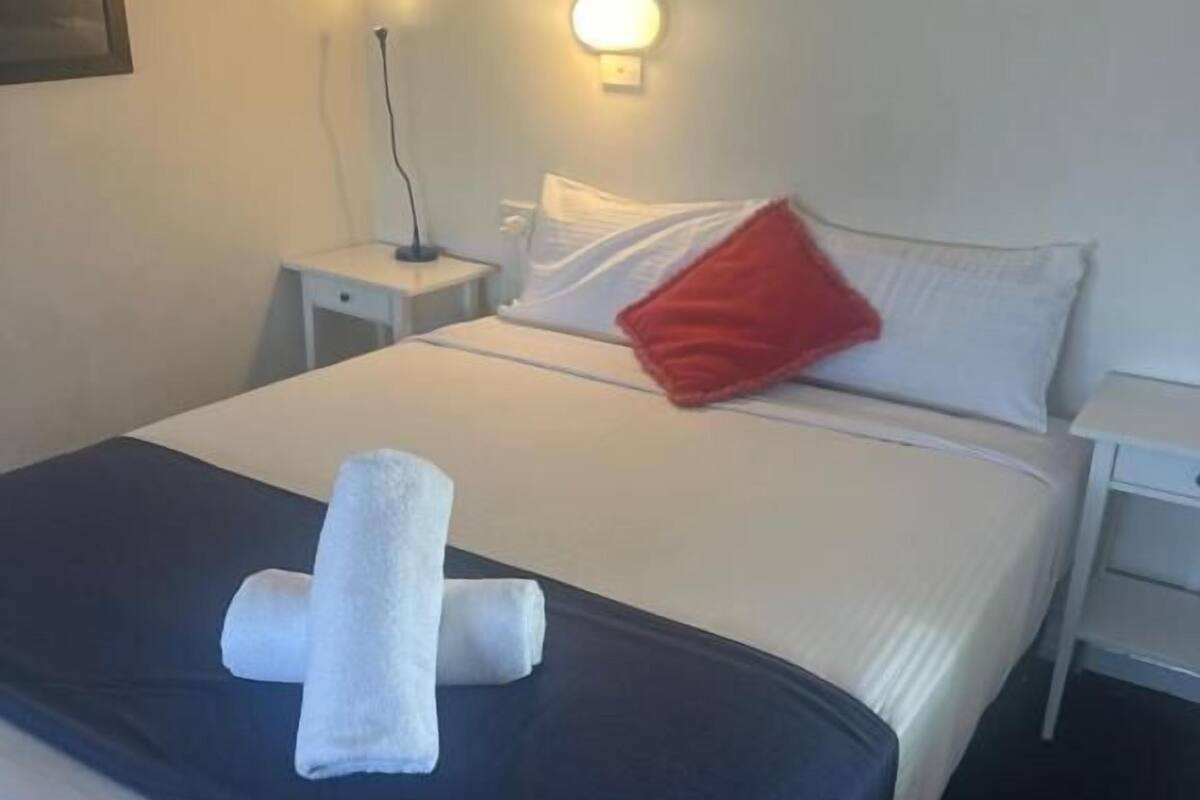 A neatly made bed with white linens, a red pillow, and a rolled white towel on the bed.