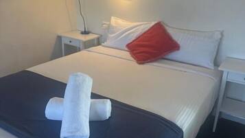 Una cama bien tendida con sábanas blancas, un almohadón rojo y una toalla blanca enrollada sobre la cama.
