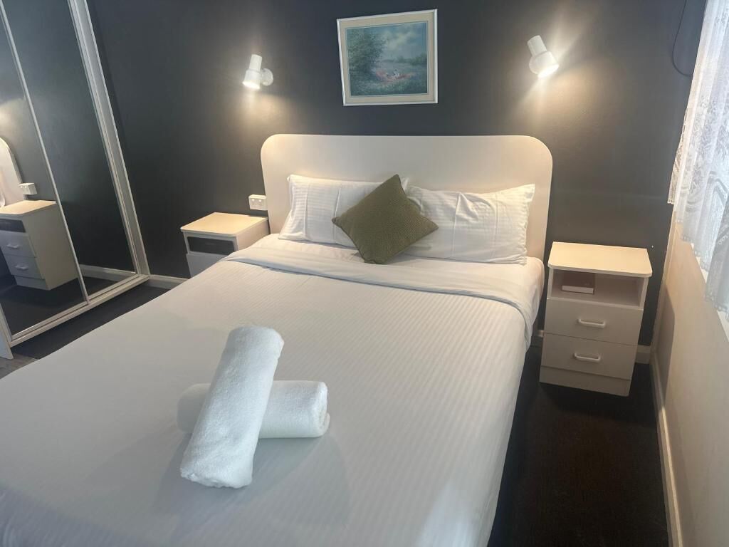 Una cama bien tendida con sábanas blancas y una toalla doblada, un almohadón verde y un cuadro en la pared.