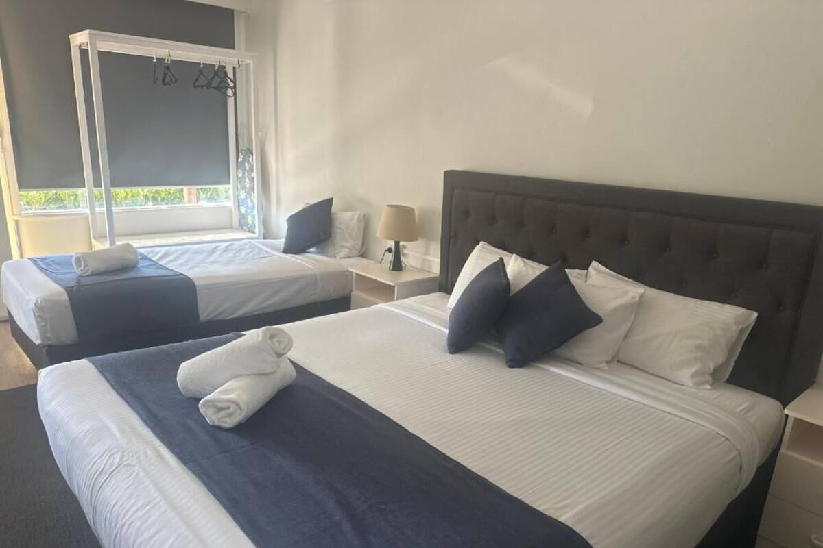 A hotel room with two beds, each with a white and blue bedspread, a dark headboard, and a nightstand with a lamp.