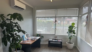 A room with a desk, two potted plants, a window with blinds, and an air conditioning unit.