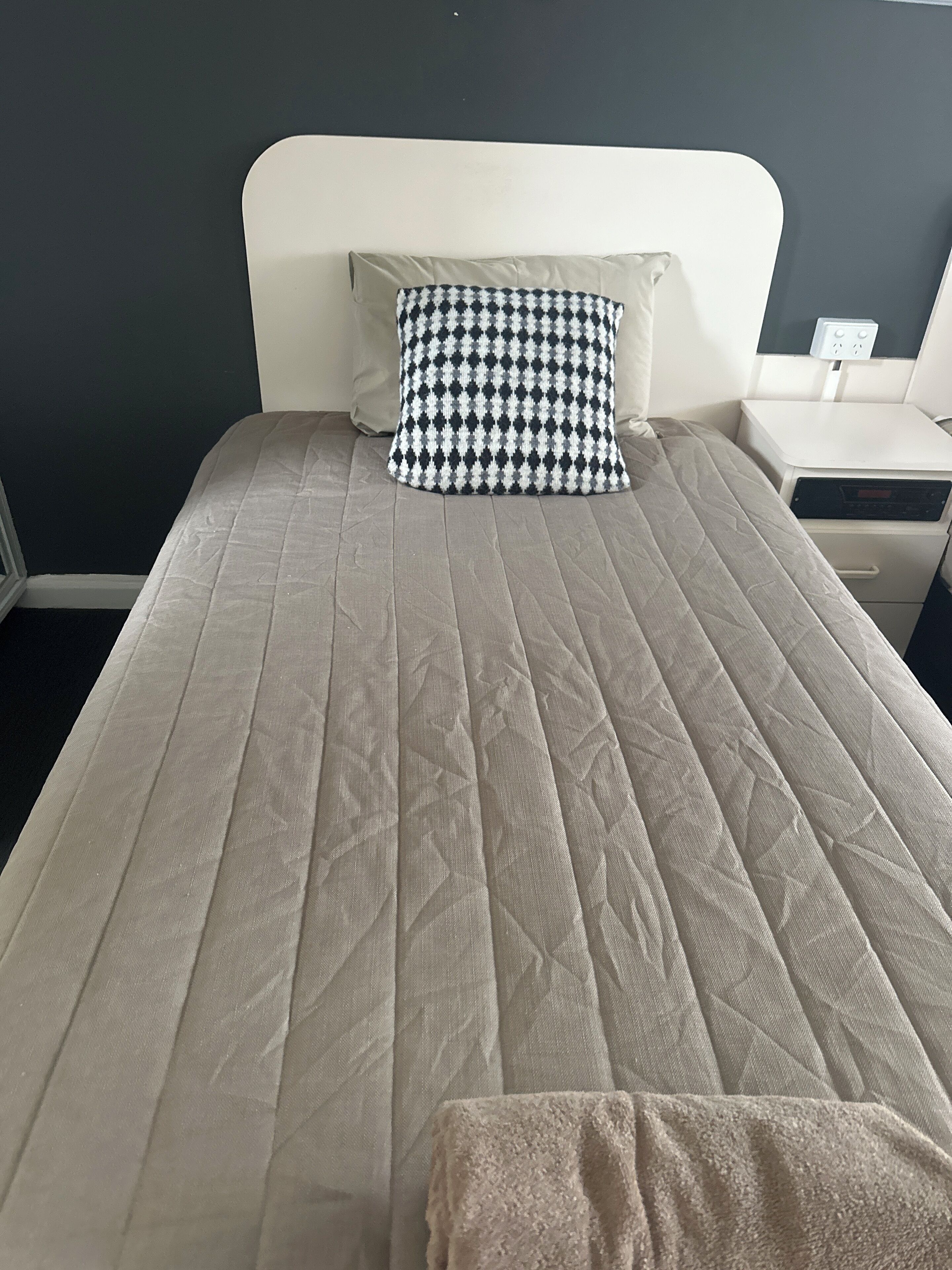 A bed with a quilted beige bedspread, a black and white checkered pillow, and a bedside table with a power outlet.