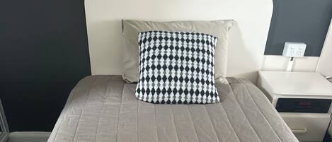 A bed with a quilted beige bedspread, a black and white checkered pillow, and a bedside table with a power outlet.