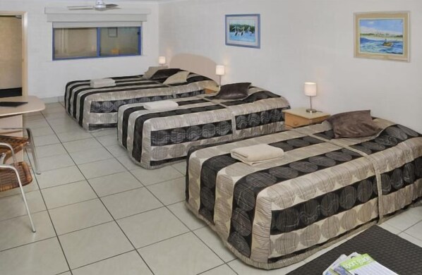 Habitación de hotel con tres camas, una mesita y sillas. Cuenta con dos ventanas y un ventilador de techo.